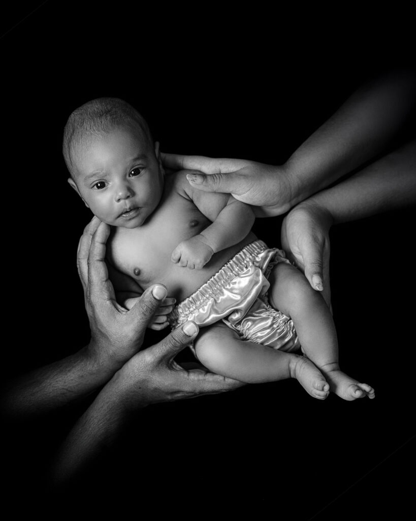 Fotografía de Recién Nacido en Clave Baja en New Jersey | Sesiones de Fotos de Bebés Artísticas Fotografía artística en clave baja de un bebé recién nacido sostenido por las manos de sus padres sobre un fondo negro. Sesión de fotos de bebés y recién nacidos en New Jersey.